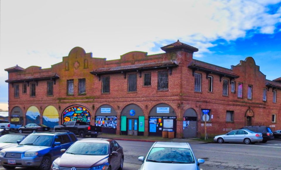 A black and white photo of a two-story brick building with seven ten-foot-tall ground-floor arches along its facade. An early 1900s-era automobile is parked in front of the building. Two men stand in the rightmost archway.