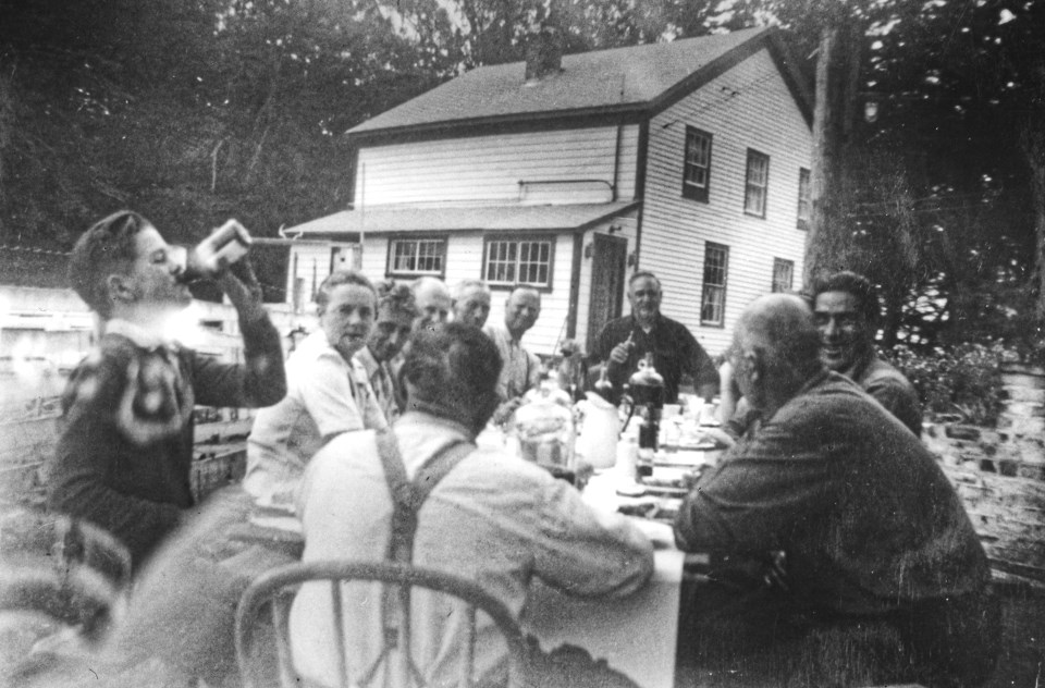 A black and white photo of men sitting outside at a table eating with a building behind them.