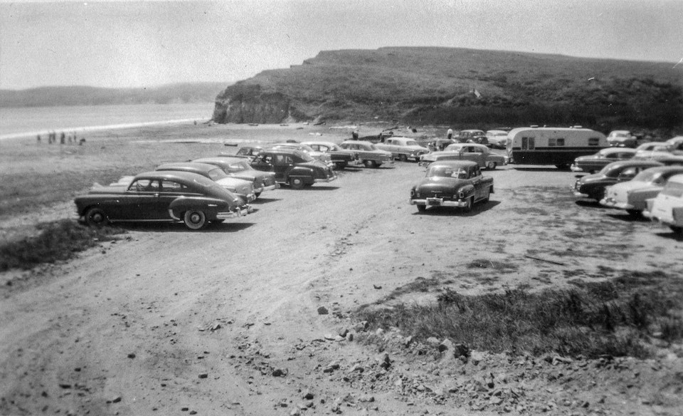 A black and white photo of cars in a parking lot, a beach, and hills, with a bay on the left..