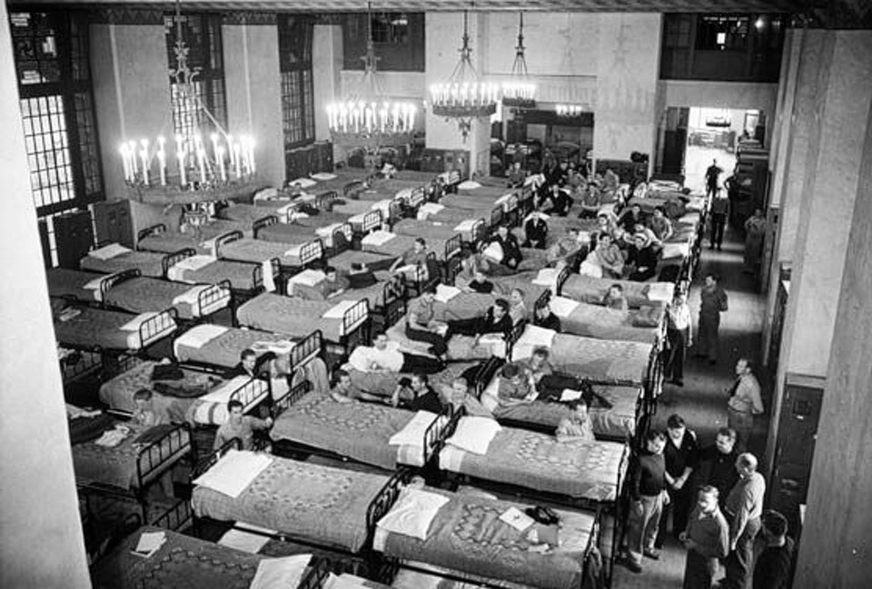 Multiple hospital beds lined up in a large room with a few people off to one side.