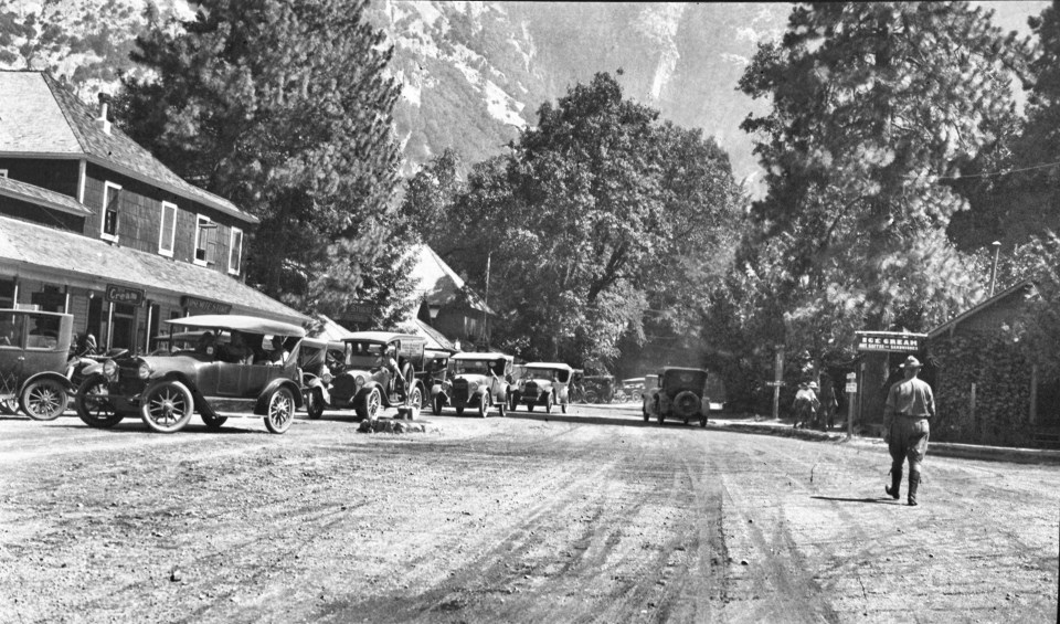 A road with a man walking, old cars parked, and a building with trees in the background.