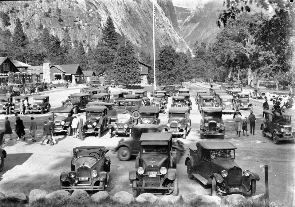 Multiple older cars in a parking lot with trees and cliffs in the background.