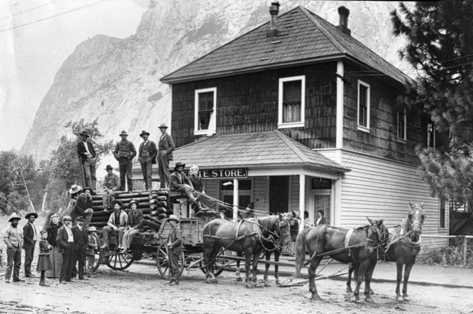 Men, a wagon with a large wooden trap, and horses in front of a two-story structure.