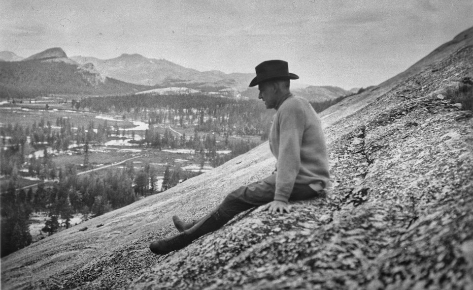 Man sitting on granite slope with meadow and mountains in background.