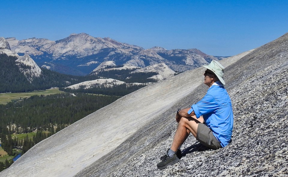 Man sitting on granite slope with meadow and mountains in background.