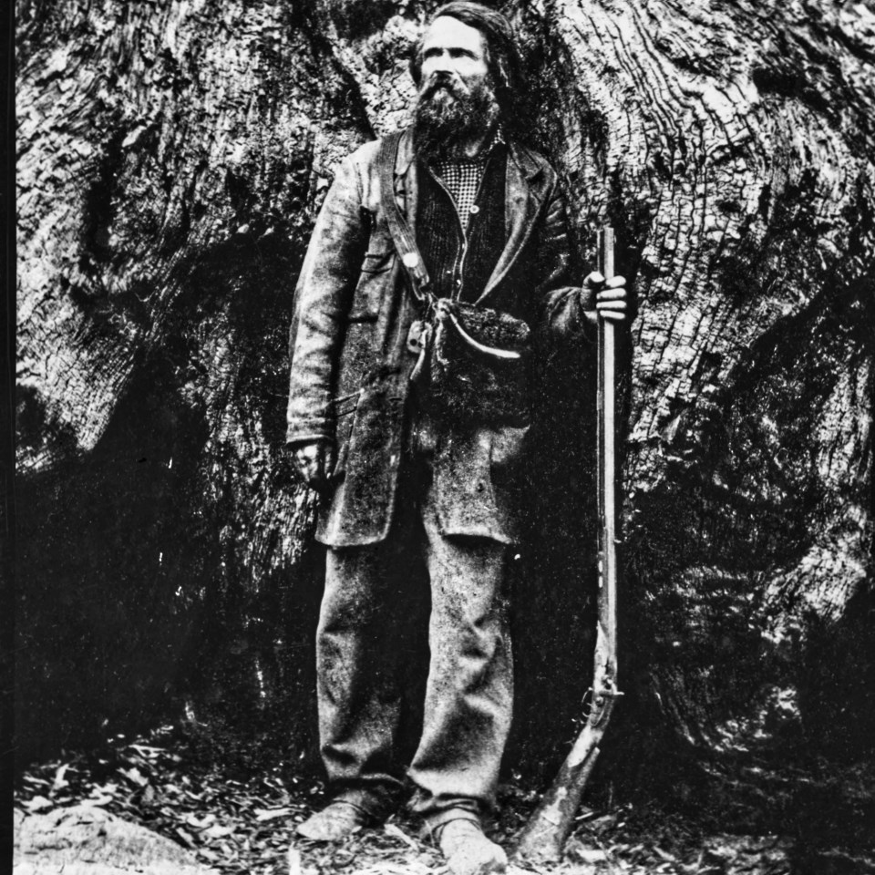 Bearded man with rifle stands at base of Giant Sequoia.