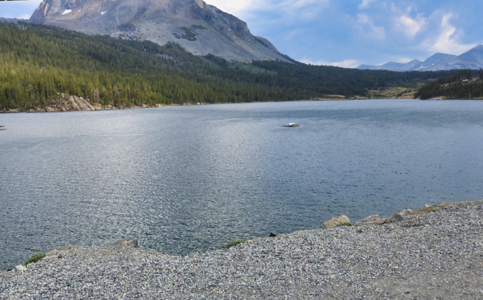 An older convertible car with several people look at a lake with mountains in background.