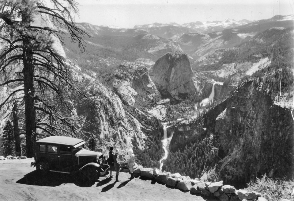 A person next to a parked older car with waterfalls and mountains in background.