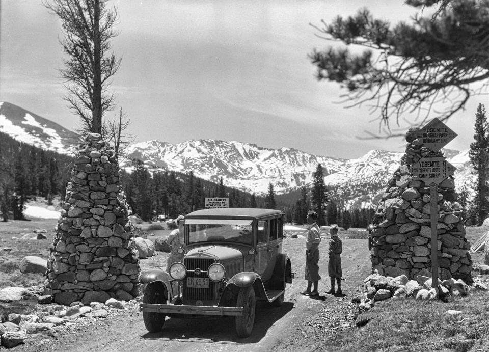 Two people stand next to an older car with two rock towers on either side of the car.