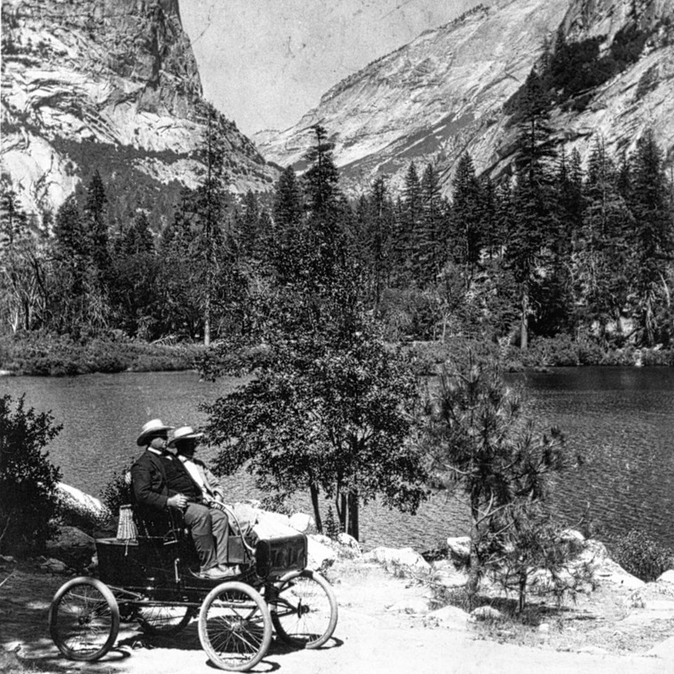 Old automobile on a trail with a lake and cliffs in background.
