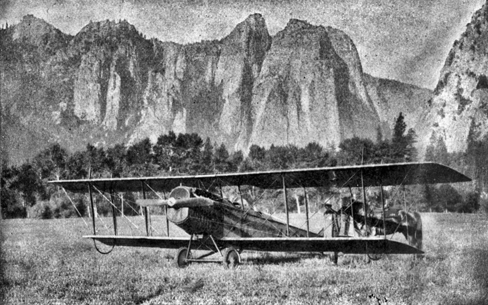 A bi-plane rests in a meadow with cliffs in the background.