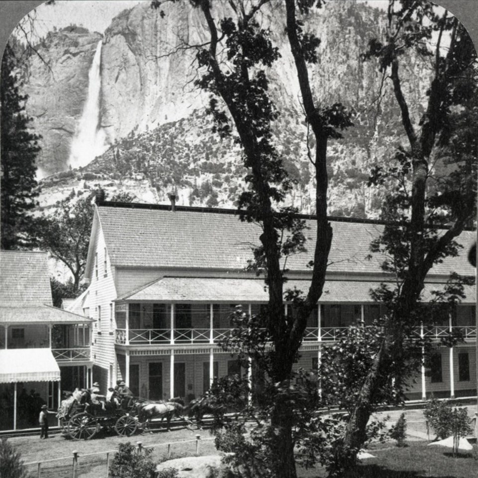 Horses and buggies in front of a building with a waterfall in the distance.