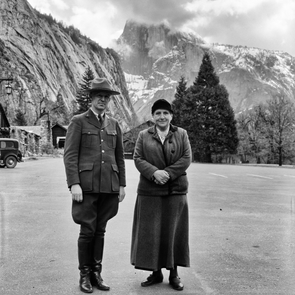 A park ranger and woman pose in a plaza with mountains in the background.
