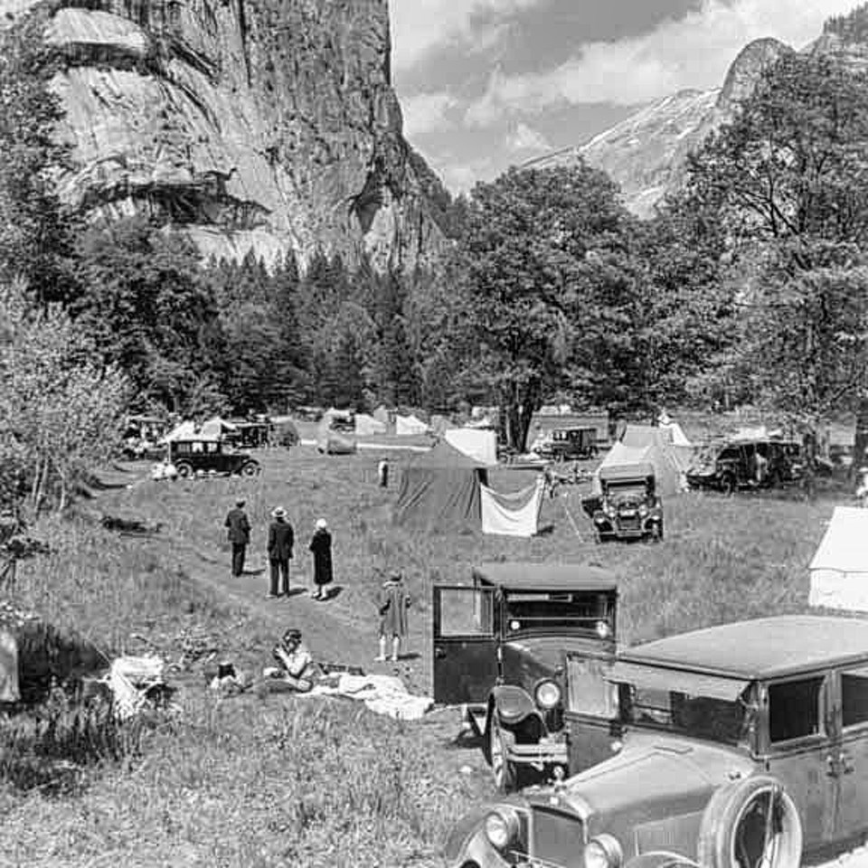 Cars, tents, and people in a meadow with trees and cliffs in background