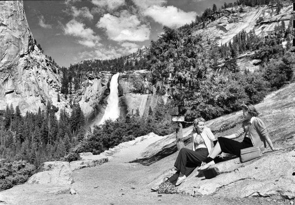 Two women sit on a granite slab next to a trail with waterfalls in the background.