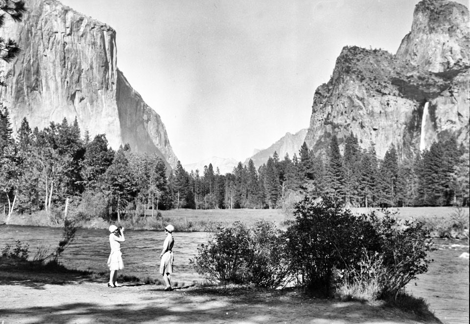 Two women on bank of river with cliffs and a waterfall in background.