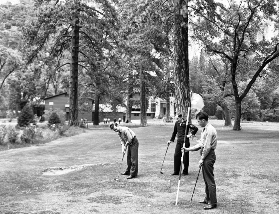 Men in naval uniform play golf on lawn with trees.