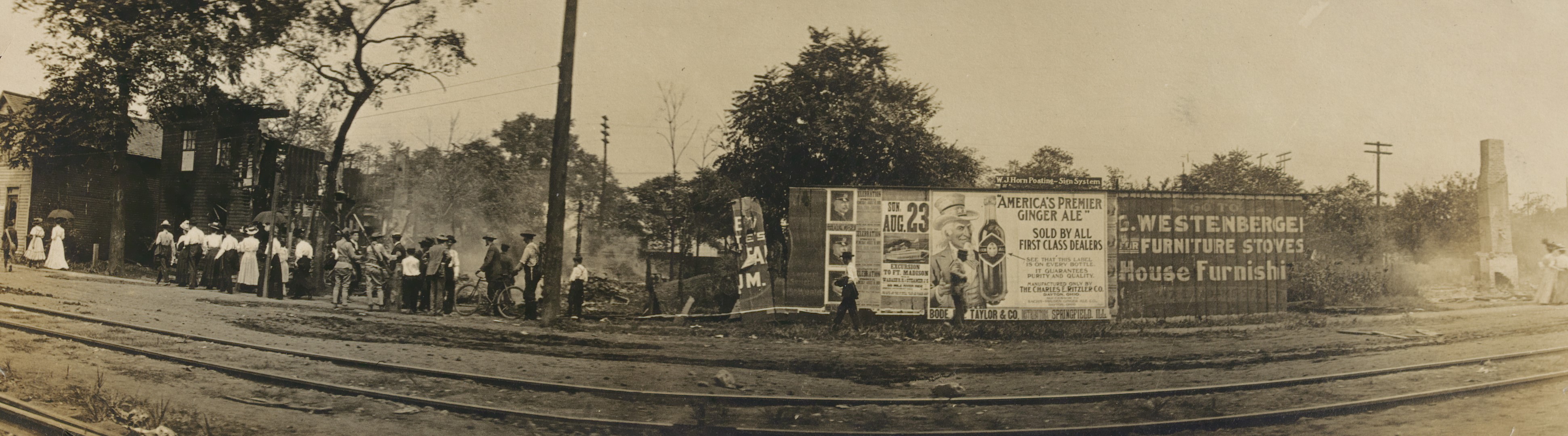 A row of smoldering and charred house frames line a city street. Onlookers and soldiers stand on the sidewalk, looking at the houses.