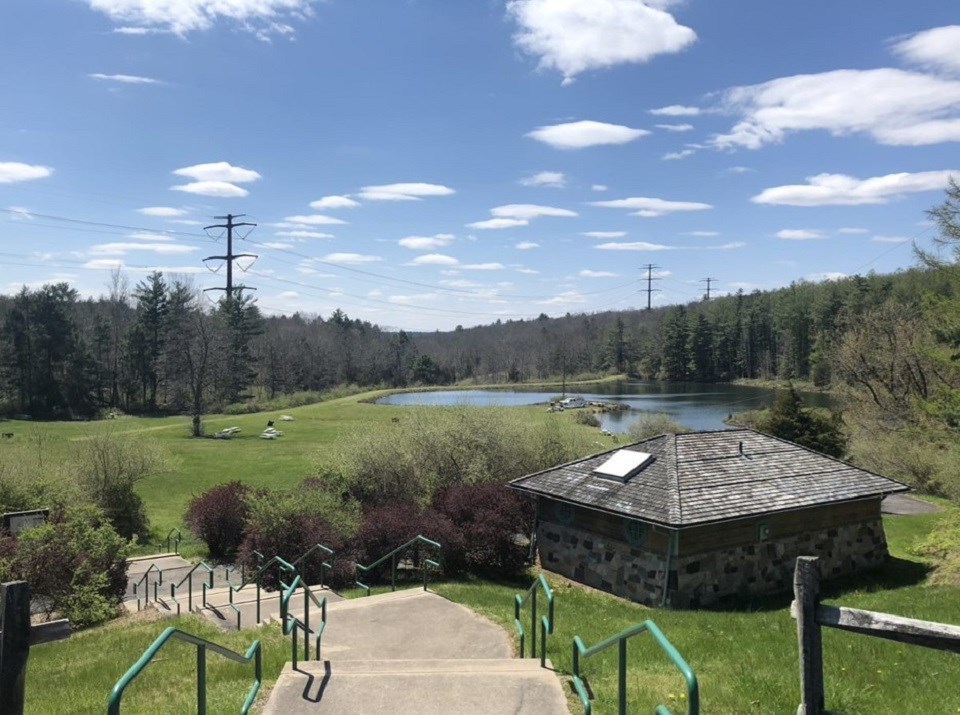 The view from the top of the stairs at Watergate Recreation Site. To the right of the staircase is a small stone restroom facility. In the distance is a large pond with trees and picnic tables surrounding it. A transmission line runs through the area.