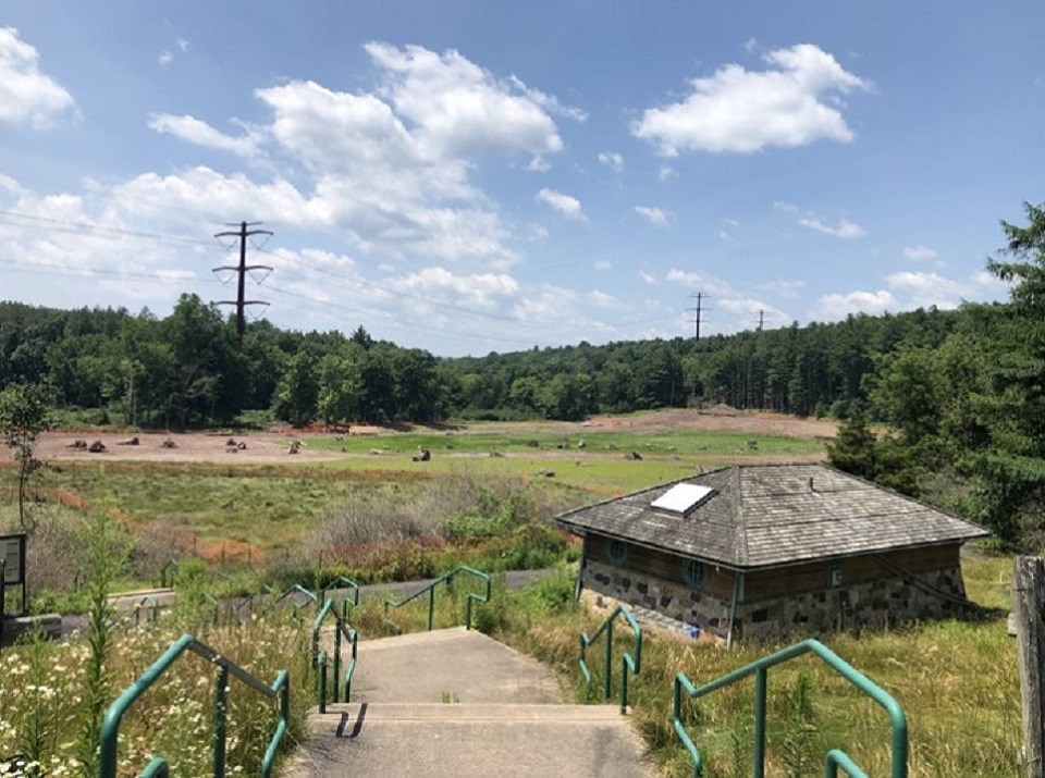 The view from the top of the stairs at Watergate Recreation Site. To the right of the staircase is a small stone restroom facility. In the distance is a large pond with trees and picnic tables surrounding it. A transmission line runs through the area.