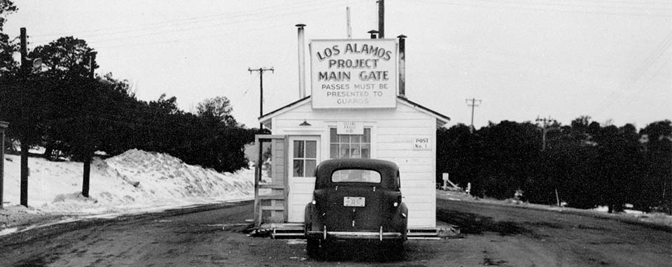A black and white photo of a white building with a big white sign that has black writing that says, "Los Alamos Project Main Gate"