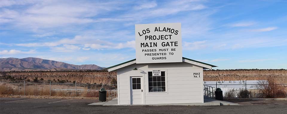 A black and white photo of a white building with a big white sign that has black writing that says, "Los Alamos Project Main Gate"