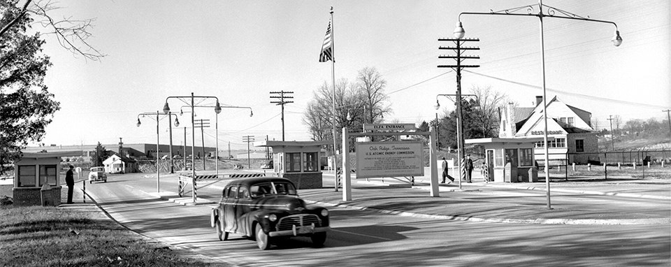 A black and white photo of a car on road going past a gate.