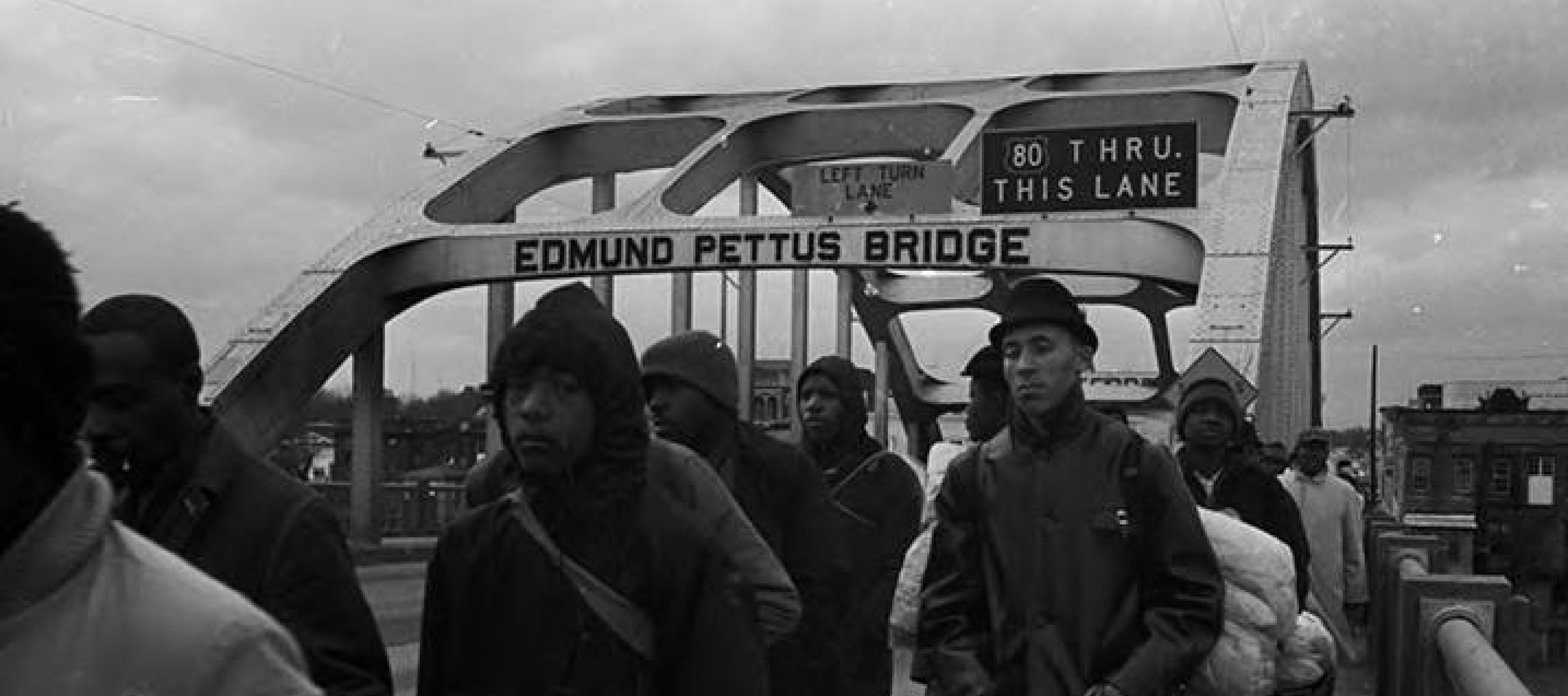 A crowd walking across a bridge, the arched steel beams overhead the people read, "Edmund Pettus Bridge,” blue sky in the background.