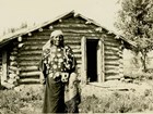 Ohchiish (Finds-Them-and-Kills-Them) standing in front of his cabin.