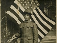 African American man wearing a World War I uniform, a United States flag is on display behind him