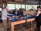 Three people stand around a large wooden table in a warehouse, looking at unrolled charts
