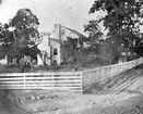 Black and white historic photo of a two-story house with a white rail fence in the yard