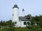 A white house with a lighthouse tower stands with a taller metal lighthouse tower behind it.