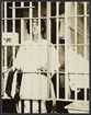 Woman standing behind bars of jail cell.