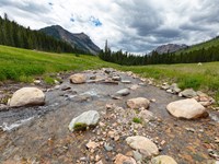 Clear, shallow, boulder-filled stream flanked by grassy meadow, forest, and mountains.