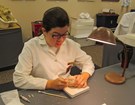 a woman in a white lab coat uses a small pick while working on a baseball-sized fossil skull.