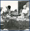 Two men kneel over a dirt pit lined with bricks. 