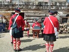 Men in bright red jackets and tartan kilts stand facing a man sitting at a table. 