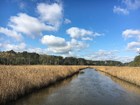 a creek cuts through golden marsh grass under a blue sky with puffy white clouds