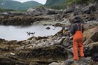 A man in orange waders operates a UAS on a rocky coast.