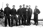 Black and white photo of seven men wearing dark clothes, standing on snow. 