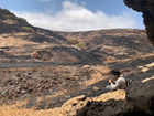 Person looking at a small object near a rock formation, with a charred landscape beyond.