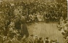 An elderly woman speaks to a crowd of well-dressed people seated on a wooden stage around her.