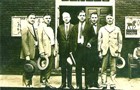 A row of men in suits and ties with hats in their hands stand on a street.
