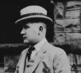 A man in a tan suit and hat stands in front of a stone wall.