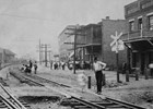 A man stands next to a railroad crossing with a row of brick buildings behind him. 
