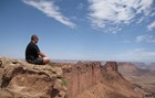 Man sitting on red sandstone rock outcrop above Farley Canyon, with blue skies and red rock canyons.