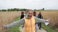 Children smile widely walking a gravel path through tall yellow grass.