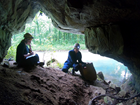 Two cavers sit at the entrance of a cave. Just outside of the cave is a clear, blue creek.