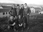 6 men pose on a hill in front of town with two-story white buildings, ocean, snowy mountains. 